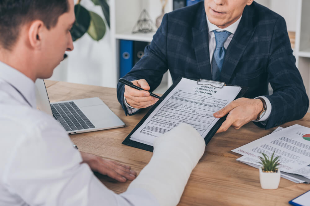 Businessman in blue jacket giving form for compensation claim to worker with broken arm at table in the office.